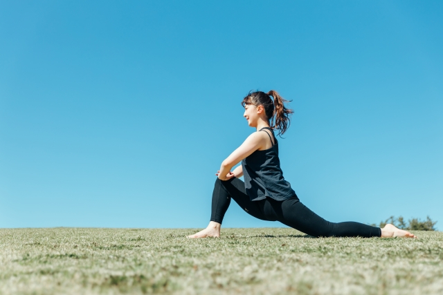 屋外で股関節をストレッチする女性。開脚ストレッチに必要な腸腰筋と大腿四頭筋を伸ばすランジポーズ（ローランジ）。青空と緑の芝生。
