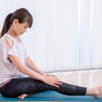 ヨガマットに座り、脚を前に伸ばして膝に手を添え、太ももの裏側やふくらはぎをストレッチするポニーテールの女性